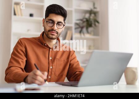 Focused Arab man in glasses using laptop and writing Stock Photo
