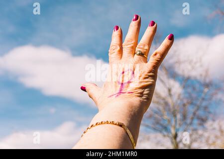 Hand of an older woman with purple painted nails with the sky in the background with the female symbol painted purple. Concept of women's day, empower Stock Photo