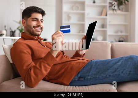 Smiling Arab guy using tablet and credit card at home Stock Photo