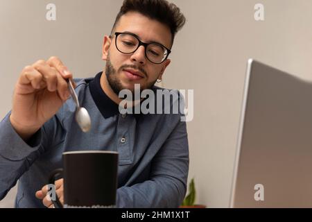 Young Latino boy stirs his coffee with a spoon while taking a break from online classes. Stock Photo