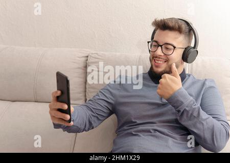 young latin guy video calling on his cell phone with his headphones on while sitting on the couch Stock Photo