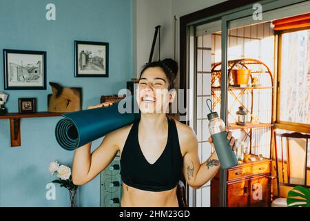 Young woman smiling and laughing to camera while holding a bottle of water and a yoga mat at a modern flat in the city during a bright day Stock Photo