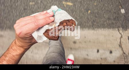 Man walking on sidewalk in the city street with bitten Chocolate Chip Cookie in his hand, point of view perspective. Stock Photo