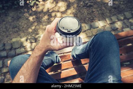 POV image of man sits on a bench and holding paper cup of coffee at a summer day in the park Stock Photo