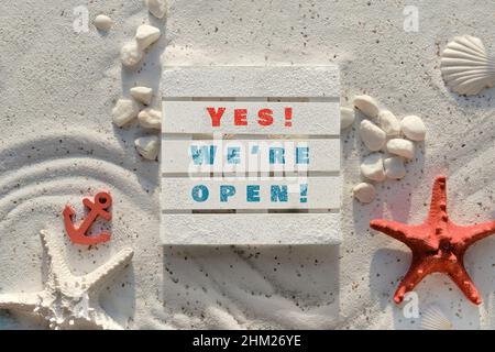 Sign yes we are open on wooden palette. Sand background with pebbles, stones, red starfish and anchor. Stock Photo