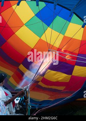 Partially-visible pilot inflating a colorful hot air balloon with a propane burner. Flame is visible. Stock Photo