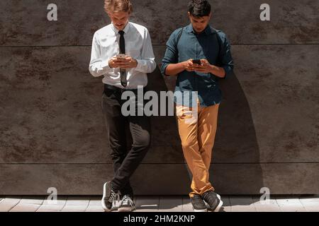 Casual multiethnic startup business men one of them is Indian using smartphone during break from work in front of grey wall outside Stock Photo