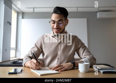 Happy smiling man sitting at desk writing in notepad listen online course Stock Photo