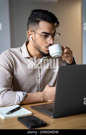 Man drinking coffee watching webinar using laptop sitting at desk in home office Stock Photo