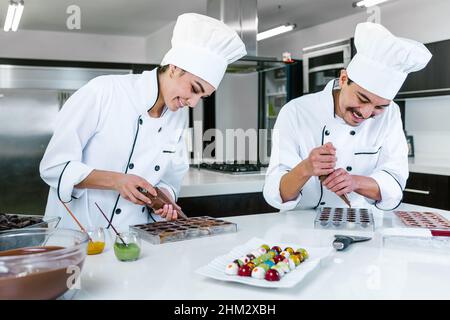 young latin couple woman and man chocolatier in chef uniform and hat preparing mexican chocolates bonbon candies at kitchen in Mexico Latin America Stock Photo