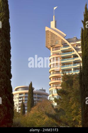 Yalta, Crimea, Russia-10.25.2019: A tall building of modern architecture with a spire, balconies behind the trees of the seaside Park in Yalta in the Stock Photo