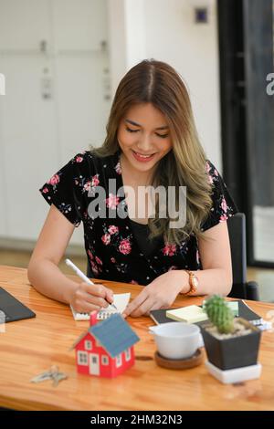 Portrait of a cheerful female house insurance agent taking list on spiral notepad at her office desk. Stock Photo