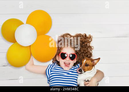 Cute girl embracing her friend jack russell dog and holding yellow balloons. Baby lying on the wooden floor with high top view. Stock Photo