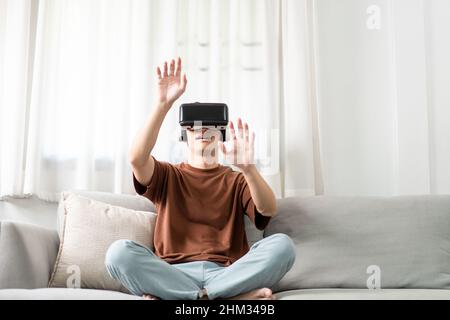 Technology Concept The man with his brown T-shirt and light blue jeans wearing a virtual reality headset while sitting on the sofa. Stock Photo