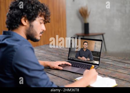 Male student studies online. Young man is watching online classes and writing a syllabus in notebook. Concept of distance study, online learning, webinars. Back view Stock Photo