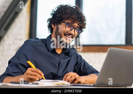 Concentrated positive Indian man in eyeglasses thinking, taking notes while sitting at the table with laptop and smartphone at home. Author writing idea for his book, study online. E-learning concept Stock Photo
