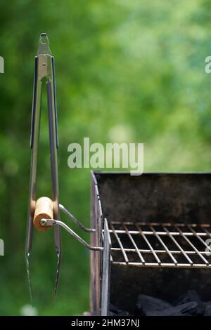 Barbecue tongs (Grillzange) on a wooden handle from a charcoal grill with dirty rust. Green nature in the background. View from the front. Stock Photo