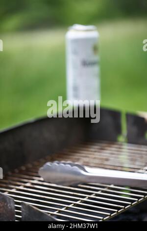 Dirty grill grate, white beer can and stainless steel grill tongs. Green nature in background. Side view. Stock Photo