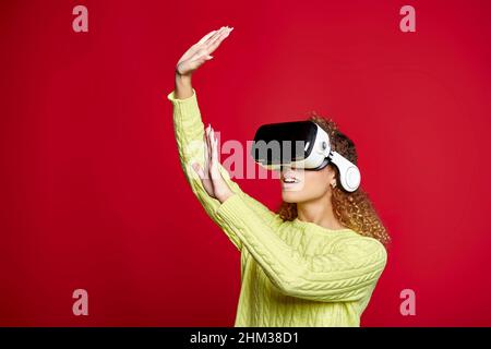 Content African American female in VR headset touching invisible object and experiencing cyberspace on red background in studio Stock Photo