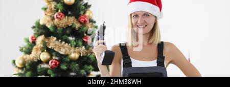 Woman builder in red santa claus hat holding drill in hands near christmas tree Stock Photo