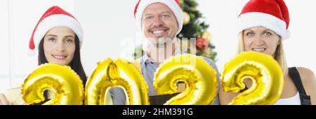 Group of repairmen holding golden helium balloons with numbers 2022 near new year tree Stock Photo