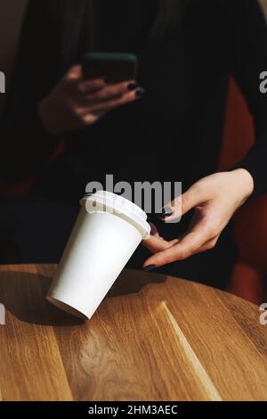 White paper cup takeaway mockup, on a table in a cafe. A woman's hand holds a cup and a smartphone. The concept of a break, rest in a cafe. Copyspace. Stock Photo
