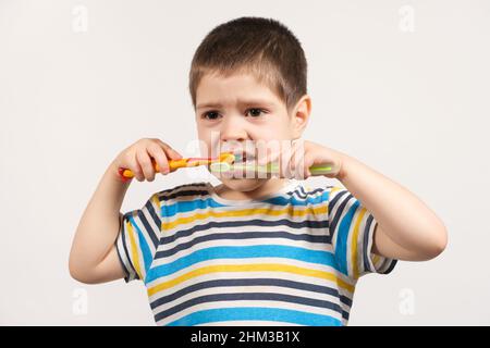 A preschooler boy brushes his teeth with two toothbrushes on a white background of isolate. Care for milk teeth in children Stock Photo