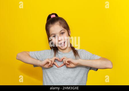 Adorable girl in gray t-shirt making a heart gesture with her fingers with a happy sincere smile isolated over yellow background. Expression of love a Stock Photo