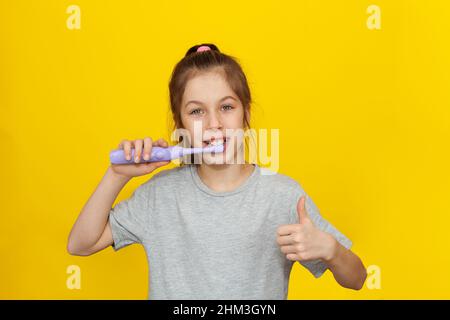 Beautiful brown-haired teenage girl holding brushing her teeth with an electric toothbrush on a yellow background, Children nutrition and oral hygiene Stock Photo