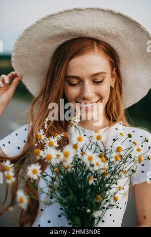Portrait of a caucasian red-haired woman in hat holding a daisies bouquet. Closeup portrait. Beautiful young girl. Chamomile flower field. Stock Photo