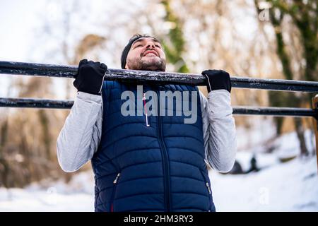 Adult man is exercising on pull-up bar in park in wintertime. Stock Photo