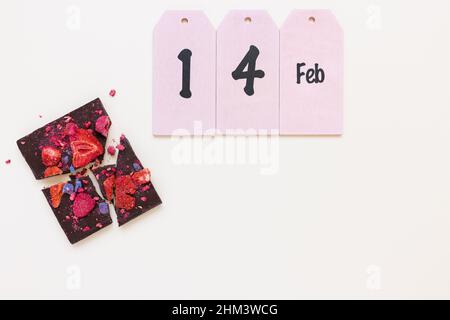 Broken handmade dark chocolate bar with dried fruit and berries and text 14 Feb on white background. Abstract Valentines Day and candy gift concept. Top view, copy space Stock Photo