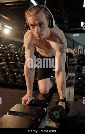 Fit concentrated young man leaning on gym bench and doing exercise for triceps with heavy Stock Photo