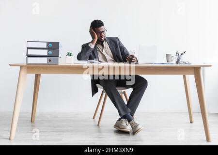 Stressed young black businessman using smartphone and laptop, sitting at desk, making error in business project Stock Photo