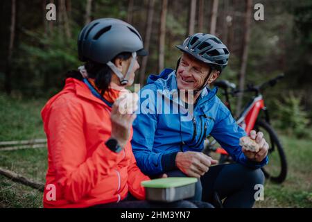 Happy senior couple bikers with eating snack outdoors in forest in autumn day. Stock Photo
