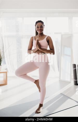 Happy young black woman in sports clothes standing in tree pose, practicing yoga on mat at home gym, full length Stock Photo