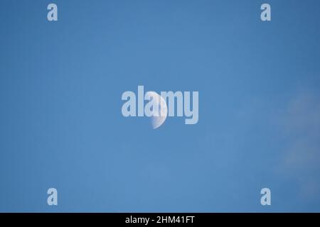 The Moon in the distance in the early morning during the First Quarter of its Lunar Phase surrounded by blue sky taken in January from the UK Stock Photo