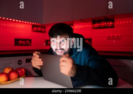 a male hacker behind a laptop in a dark room holds his hands on the computer display Stock Photo