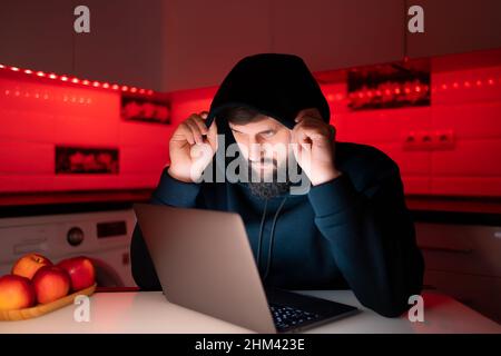 a hacker in a black hoodie with a hood on his head is sitting at a laptop and holding the hood Stock Photo