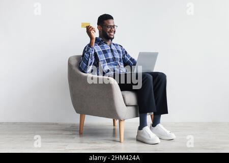 Online shopping. Young black man sitting in armchair with laptop, using credit card to buy goods on web Stock Photo