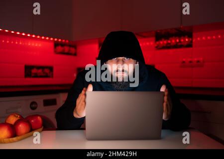 a hacker in a black hoodie with a hood on his head is sitting at a laptop holding its lid and looking suspiciously Stock Photo