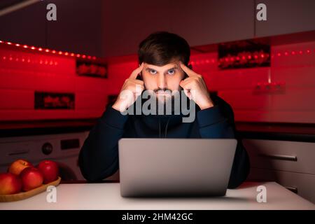 a hacker in a black hoodie is sitting at his laptop thinking Stock Photo