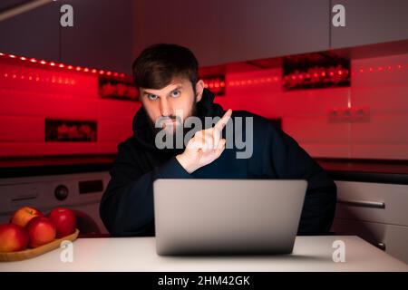 a hacker in a black hoodie is sitting at a laptop pointing his fingers to the side Stock Photo