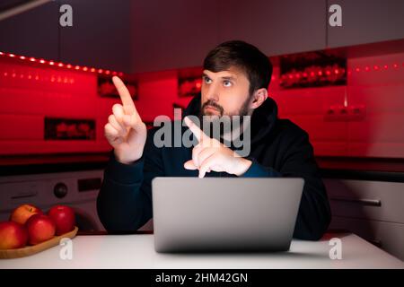 a hacker in a black hoodie is sitting at a laptop pointing his fingers to the side Stock Photo