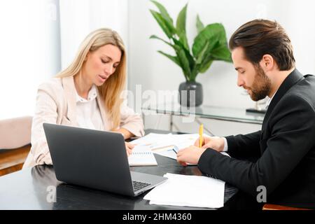Working process of two colleagues in busy atmosphere. Attractive young woman sitting with guy, they attentively writing notes. Team in office concept Stock Photo