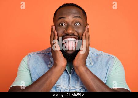 Photo of shocked cute afro guy wear denim vest arms cheeks open mouth isolated orange color background Stock Photo