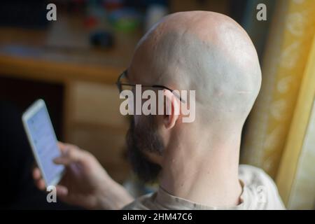 The bald man looks at the phone. The guy is looking for information in his smartphone. The guy's shaved head. Middle aged man. The guy holds a mobile Stock Photo