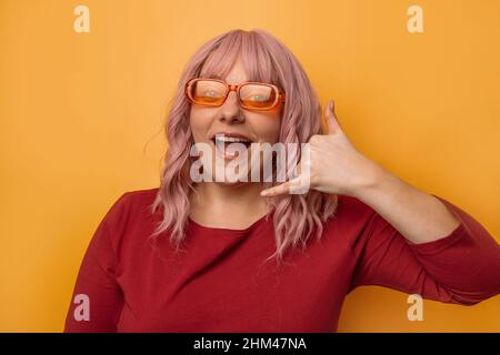 Beautiful smiling girl showing call me, smiling camera have fun shows call me back sign with hand near ear gesture isolated yellow background. Stock Photo