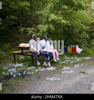 Two adults and child in gas masks sitting on wooden bench in the middle of forest. Plastic bottles scattered around on grass and road. On backdrop containers, filled by garbage, bottles, plastic bag. Stock Photo