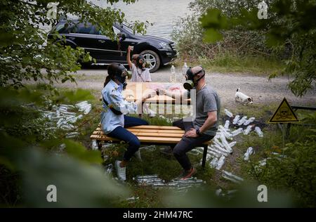 Man and woman in gas masks sitting on bench and toasting with plastic cups while spending time with daughter in roadside forest with trash plastic bottles and poison toxic sign. Ecology concept. Stock Photo